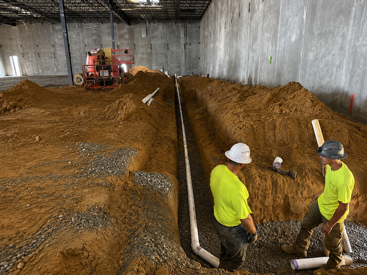 Two Webster & Sons plumbers installing PVC sanitary drain piping in underground trench inside BJ's Wholesale Club building shell, Selma NC